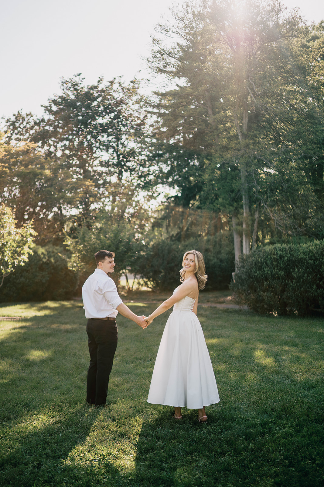 groom and bride-to-be hold hand walking in the garden while looking back wearing your white formal outfit at Morven Park during their Engagement session